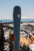 A person in a tan jacket and brown glove holds an upright Icelantic Skis Torrent 88, with snow-covered mountains and trees in the background under a clear sky.