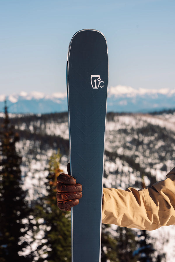 A person in a tan jacket and brown glove holds an upright Icelantic Skis Torrent 88, with snow-covered mountains and trees in the background under a clear sky.