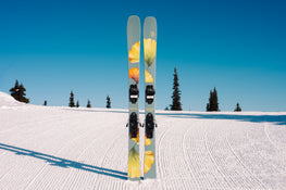 A pair of Icelantic Skis Tempest 100 all-mountain skis with yellow and green flower designs stand upright on a groomed snowy slope, featuring a poplar and birch wood core for stability and responsiveness. In the background, clear blue sky stretches above distant pine trees.