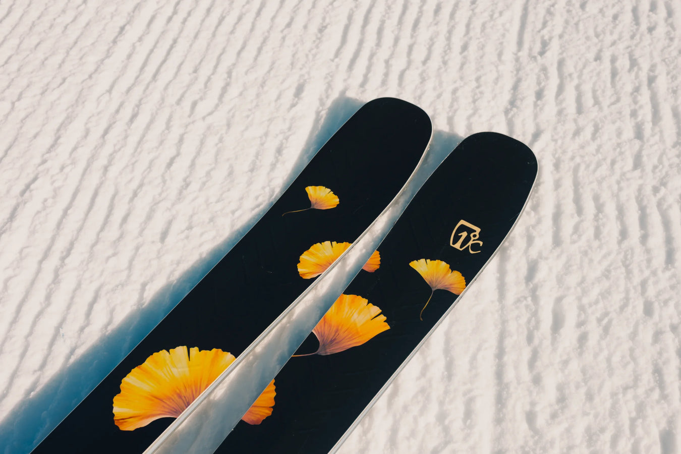A close-up of two Icelantic Skis Tempest 88 black skis featuring orange flower designs, resting on textured, groomed snow.