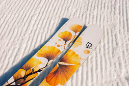 A close-up of two Icelantic Skis Tempest 94 skis with yellow leaf designs resting on groomed, textured snow.