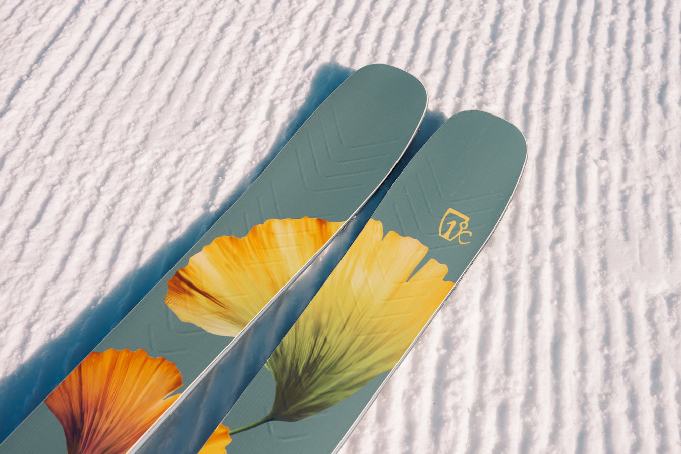 A close-up of two Icelantic Skis Tempest 100 all-mountain skis resting on snow, each featuring bold yellow-orange flower graphics and the Icelantic logo on the right ski tip. The snow shows neat, parallel grooves.