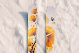 Two Icelantic Skis Tempest 94 skis, featuring white topsheets with orange and yellow floral designs, rest on the snow and cast soft shadows.