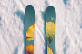 A pair of Icelantic Skis Tempest 100 all-mountain skis, featuring yellow and orange leaf designs, rest on fresh white snow, viewed from above.