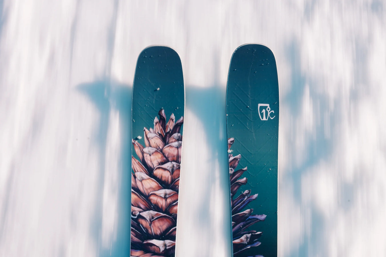 A close-up view of two Icelantic Skis Torrent 102 skis featuring a pine cone design gliding over snow, casting shadows on the snowy surface below.