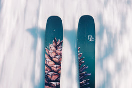A close-up view of two Icelantic Skis Torrent 102 skis featuring a pine cone design gliding over snow, casting shadows on the snowy surface below.