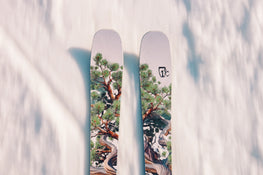 A close-up top view of two Icelantic Skis Torrent 96 skis, featuring a painted design of green pine trees and branches, gliding over smooth, white snow.