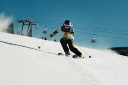 Wearing a helmet and goggles, a skier carves down a snowy slope on a sunny day, riding the Icelantic Skis Tempest 100 all-mountain skis with a poplar and birch wood core, while ski lifts and trees line the background.