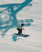 Wearing a helmet and goggles, a skier carves tracks down a snowy slope on their Icelantic Skis Tempest 100 all-mountain skis, while the large shadow of a ski lift structure stretches across the snow in the background.