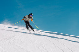 A skier in a gray jacket and black helmet is skiing downhill on a snowy slope under a clear blue sky, using yellow ski poles and carving confidently on the Icelantic Skis Tempest 100 all-mountain skis, engineered with a poplar and birch wood core.