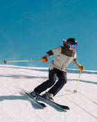 A person in a helmet and goggles skillfully skis downhill on a snowy slope under a clear blue sky, holding ski poles and wearing a beige jacket with black pants. They expertly navigate the terrain on Icelantic Skis Tempest 100 all-mountain skis, designed with a poplar and birch wood core for enhanced stability and performance.