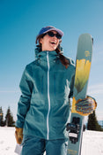A woman in a blue ski jacket and cap smiles as she holds her Icelantic Skis Tempest 100 all-mountain skis—crafted with a poplar and birch wood core and decorated with a yellow flower—while standing on snow with trees and a blue sky in the background.