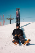 A person in black winter gear sits on snow with Icelantic Skis’ Torrent 102 skis planted upright behind them, creating a playful effect. Red ski poles and a ski lift are visible in the background under a clear blue sky.