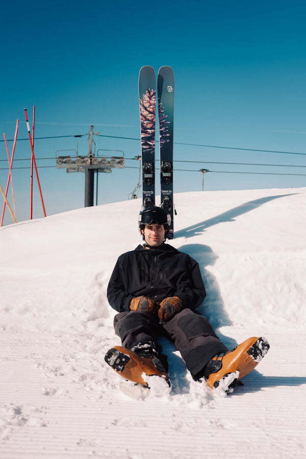 A person in black winter gear sits on snow with Icelantic Skis’ Torrent 102 skis planted upright behind them, creating a playful effect. Red ski poles and a ski lift are visible in the background under a clear blue sky.