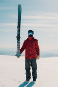 A skier in a red jacket and helmet stands on a snowy mountain holding an Icelantic Skis Torrent 102 vertically, with bright sunlight and a clear sky in the background.