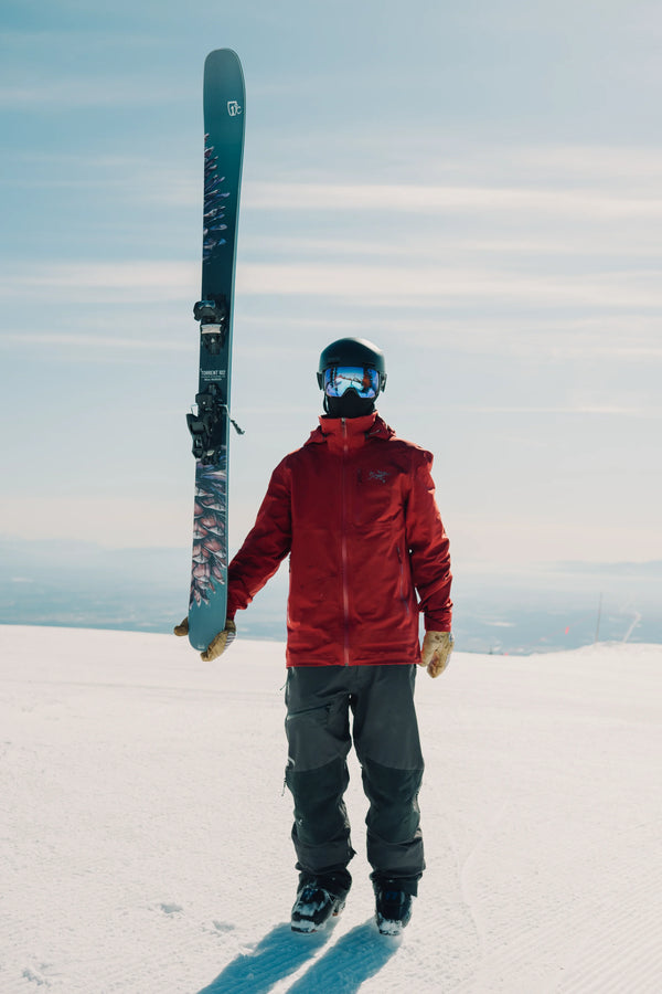 A skier in a red jacket and helmet stands on a snowy mountain holding an Icelantic Skis Torrent 102 vertically, with bright sunlight and a clear sky in the background.