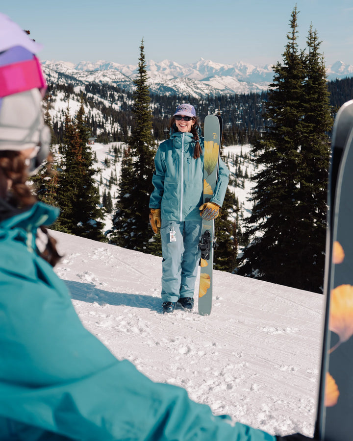 A person in blue ski gear stands smiling on a snowy mountain slope, holding a pair of Icelantic Skis Tempest 100 all-mountain skis upright, with trees and snow-covered peaks in the background. Another skier is in the foreground, partially visible.