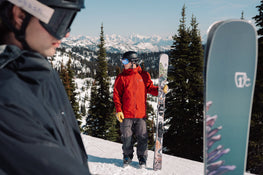 Wearing a red jacket and ski gear, a person stands on a snowy mountain holding Icelantic Skis’ Torrent 96 skis, with another skier in the foreground and trees and mountains visible in the background.