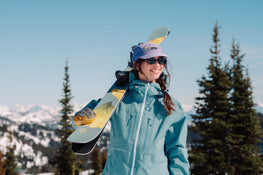 A person in a blue jacket and purple hat smiles while holding Icelantic Skis Tempest 100 all-mountain skis on their shoulder, standing outdoors with snow and pine trees in the background on a sunny day.