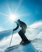 A skier carves down a snowy slope beneath a bright sun, fresh powder spraying around them as they ride the Torrent 96 from Icelantic Skis, framed by a clear blue sky.