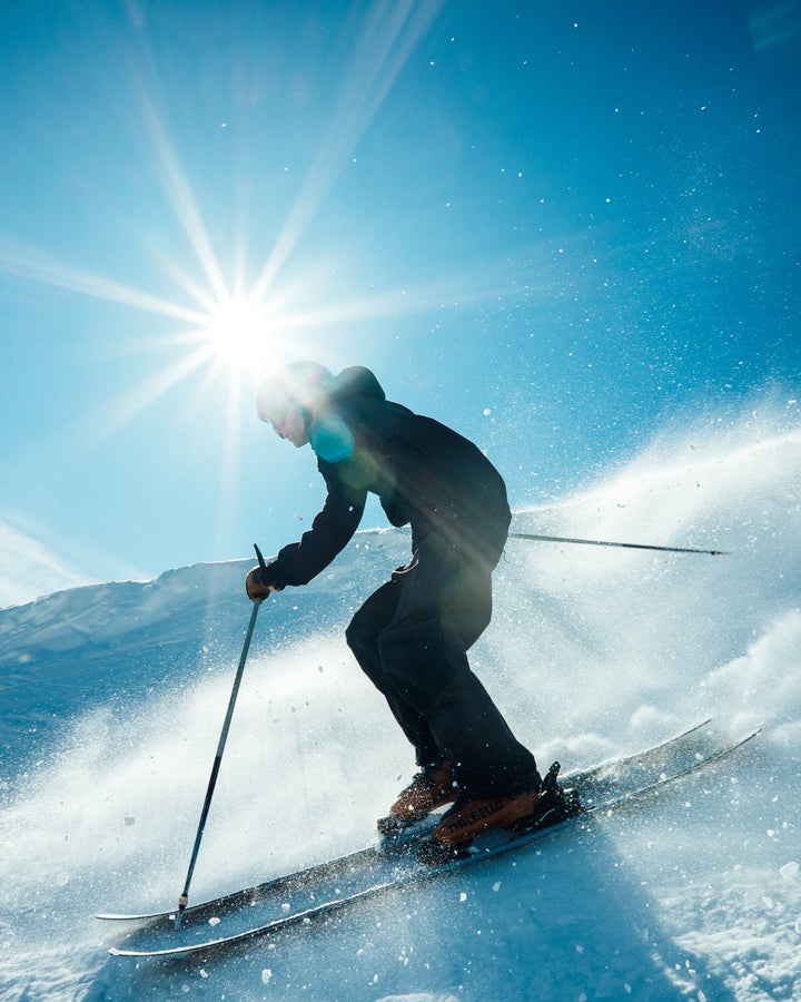 A skier carves down a snowy slope beneath a bright sun, fresh powder spraying around them as they ride the Torrent 96 from Icelantic Skis, framed by a clear blue sky.