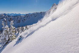 A skier carves down a steep, snowy mountain slope on Icelantic Skis’ Nomad 112, sending up a spray of powder beneath a clear blue sky, with pine trees and rugged peaks rising in the background.