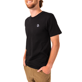 A man stands against a white background with his hands in his pockets, smiling, dressed in tan pants and the Icelantic Embroidered One Degree Tee in black. This tee features a small white embroidered logo on the chest and offers both comfort and style with its unisex fit and organic cotton fabric.