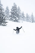 Clad in dark winter gear, a skier glides through deep snow on Icelantic Skis’ Nomad 112, surrounded by snow-laden trees during heavy snowfall.