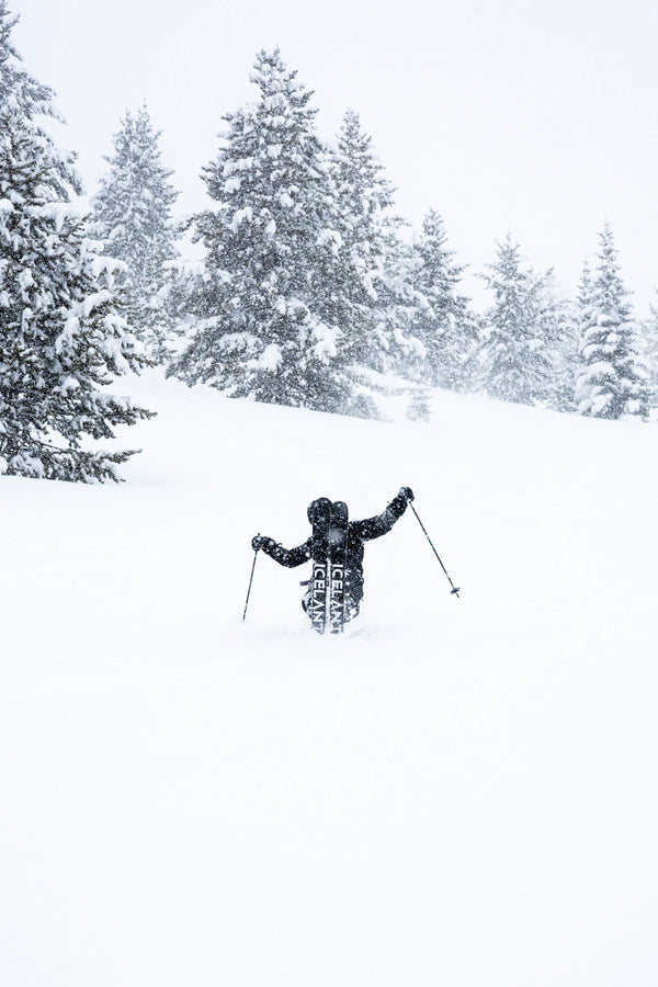 Clad in dark winter gear, a skier glides through deep snow on Icelantic Skis’ Nomad 112, surrounded by snow-laden trees during heavy snowfall.