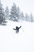 Clad in black gear, a skier carves through deep snow on a snowy slope atop Icelantic Skis’ Nomad 112 Whiteout, surrounded by snow-covered evergreens and gently falling snow.