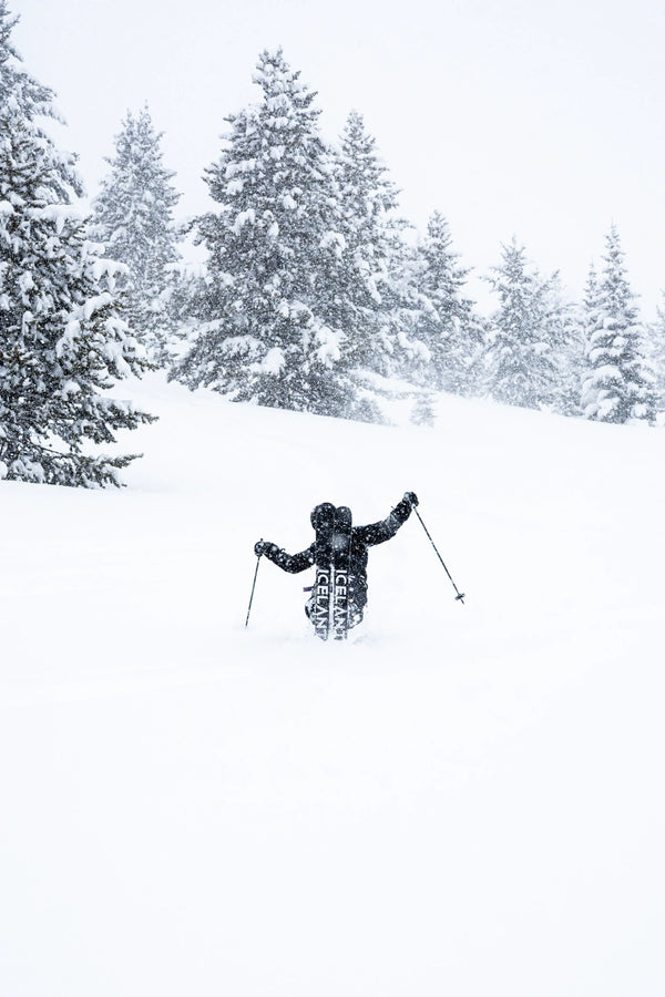 Clad in black gear, a skier carves through deep snow on a snowy slope atop Icelantic Skis’ Nomad 112 Whiteout, surrounded by snow-covered evergreens and gently falling snow.