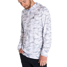 A bearded man stands against a white background, wearing the Icelantic UV Sun Hoodie - Snow Camo. This long-sleeved, UPF 50 performance hoodie features a white and light grey digital camouflage print with a small black logo on the chest, and is designed for quick-dry comfort as he casually rests his hands in his pockets.