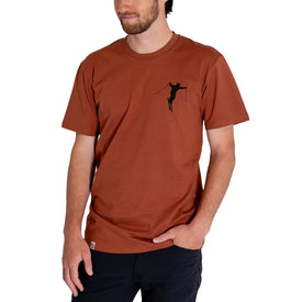 A man wearing the Icelantic Cowboy Skier Tee from the Into the West summer tees collection, featuring a small black cowboy skier graphic on the upper left chest. Crafted from 100% combed cotton, this ski-themed t-shirt in rust color is paired with black pants.