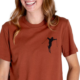 A smiling person wearing an Icelantic Cowboy Skier Tee in rust, crafted from 100% combed cotton, featuring a black silhouette of a skier on the left chest.