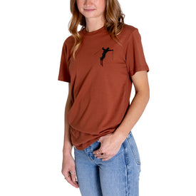 A woman wearing an Icelantic Cowboy Skier Tee in rust, crafted from 100% combed cotton and featuring a small black skier graphic on the chest, paired with light blue jeans, standing against a white background.