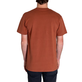 A person stands with their back to the camera, wearing Icelantic's Cowboy Skier Tee—a short-sleeve, rust-colored shirt crafted from 100% combed cotton—paired with dark pants against a white background. This look perfectly channels the laid-back style of Into the West summer tees.