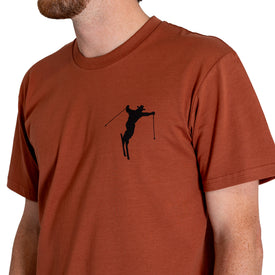 A person wearing the Icelantic Cowboy Skier Tee—a rust-colored t-shirt featuring a small black silhouette of a skier holding poles on the left chest. Crafted from 100% combed cotton, only the upper body and part of the face are visible.