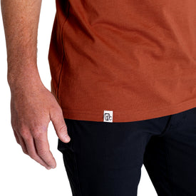 A person wearing the Icelantic Cowboy Skier Tee in rust-colored 100% combed cotton and black pants stands with their left arm relaxed by their side. A small white tag with black lettering is visible on the T-shirt’s hem.