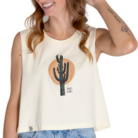 A woman smiles, wearing the Icelantic Cactus Cropped Tank in linen—a sleeveless white women’s cotton top featuring a cactus graphic and the words "Into the West"—paired with denim shorts. Perfect summer gear, her inner arm tattoo is visible.