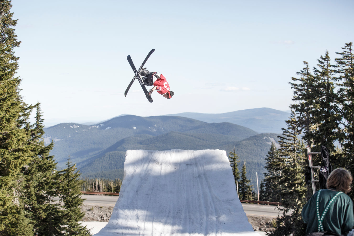 An athlete equipped with Icelantic Skis’ Nomad 100 in a red jacket executes an aerial trick above a snowy ramp, set against pine trees and distant mountains beneath a clear sky.