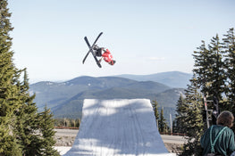 An athlete equipped with Icelantic Skis’ Nomad 100 in a red jacket executes an aerial trick above a snowy ramp, set against pine trees and distant mountains beneath a clear sky.