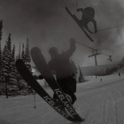 A skier performs a trick mid-air on a snowy slope lined with pine trees, riding the Icelantic Skis Nomad 100 Whiteout, while another skier soars in the background and a ski lift filled with riders appears in the distance. The scene is captured in black and white.