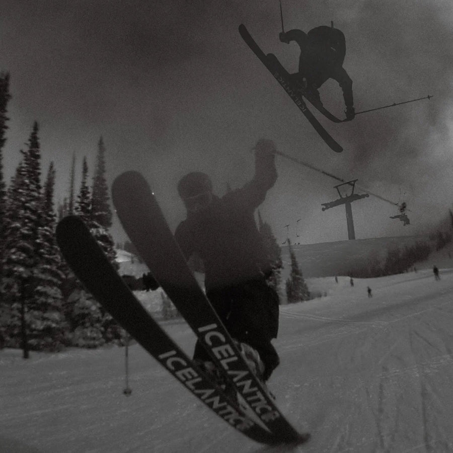 A skier performs a trick mid-air on a snowy slope lined with pine trees, riding the Icelantic Skis Nomad 100 Whiteout, while another skier soars in the background and a ski lift filled with riders appears in the distance. The scene is captured in black and white.