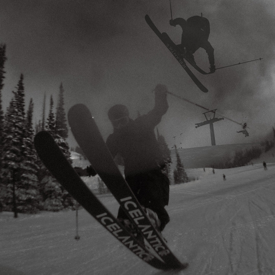 A skier performs a trick on a snowy slope using Icelantic Skis’ Nomad 100 skis. Another skier jumps in the background near a ski lift, with snow-covered trees on the side. The image is in black and white.