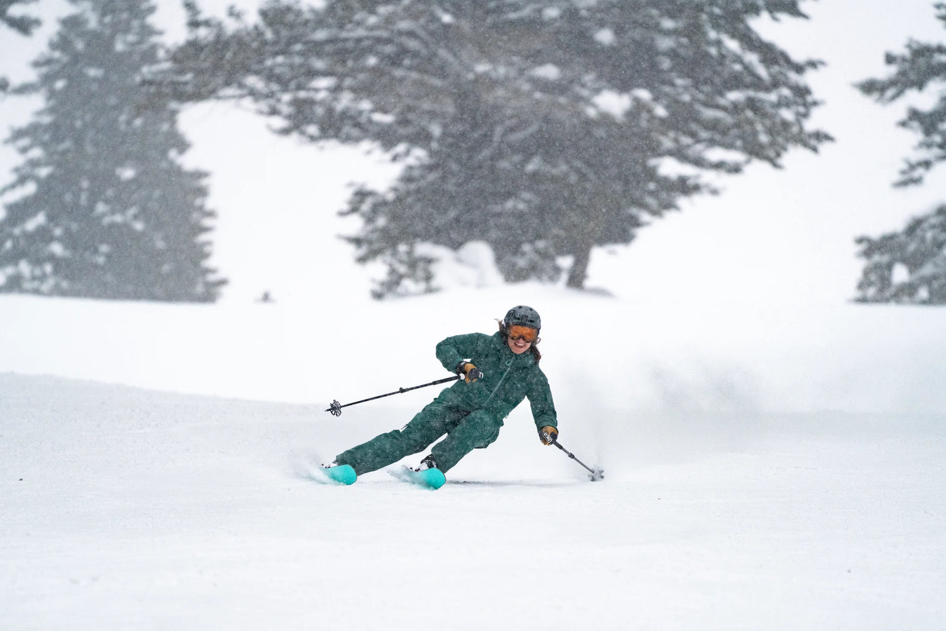 Wearing green ski gear and a helmet, a skier carves downhill on a snowy slope with the Icelantic Skis Maiden 94, kicking up a spray of snow as snow-covered trees fill the background.