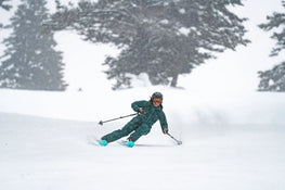 Wearing green ski gear and a helmet, a skier carves downhill on a snowy slope with the Icelantic Skis Maiden 94, kicking up a spray of snow as snow-covered trees fill the background.