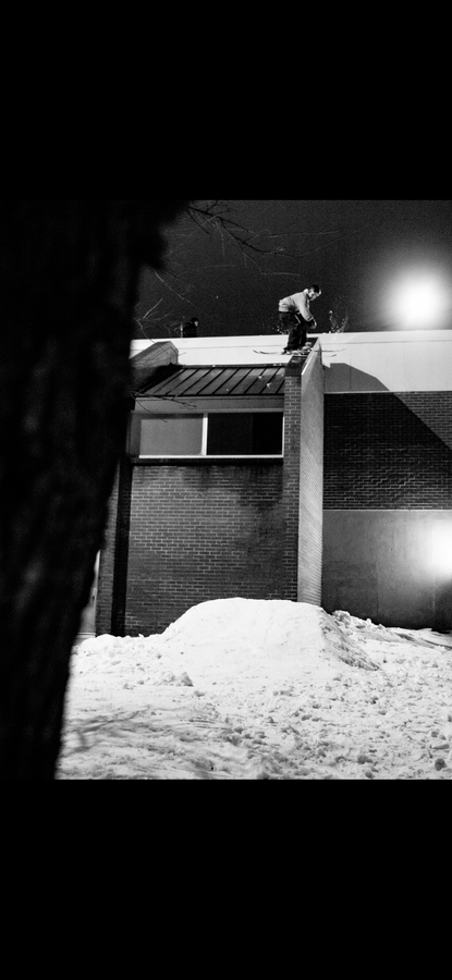 A snowboarder, riding Icelantic Skis’ Nomad 94 Whiteout, prepares to jump off a building roof onto the snowy ground at night; the black and white scene features a tree trunk in the foreground.
