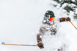 Riding on Icelantic Skis’ Nomad 112, a skier in brown gear and a black helmet carves through deep powder, sending up sprays of snow that partially obscure their face and body.