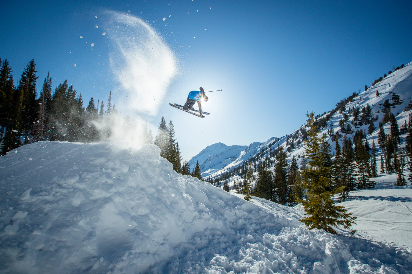 An athlete launches into the air on a snowy slope, powder flying as they ride the Nomad 106 skis from Icelantic Skis. Snow-covered mountains and pine trees stretch out beneath a clear blue sky.