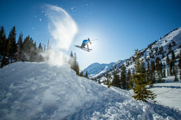 An athlete launches into the air on a snowy slope, powder flying as they ride the Nomad 106 skis from Icelantic Skis. Snow-covered mountains and pine trees stretch out beneath a clear blue sky.
